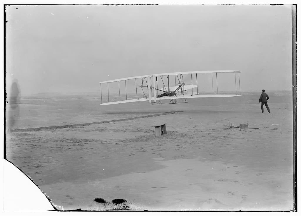 Orville Wright taking off in the Wright Flyer as Wilbur Wright runs alongside